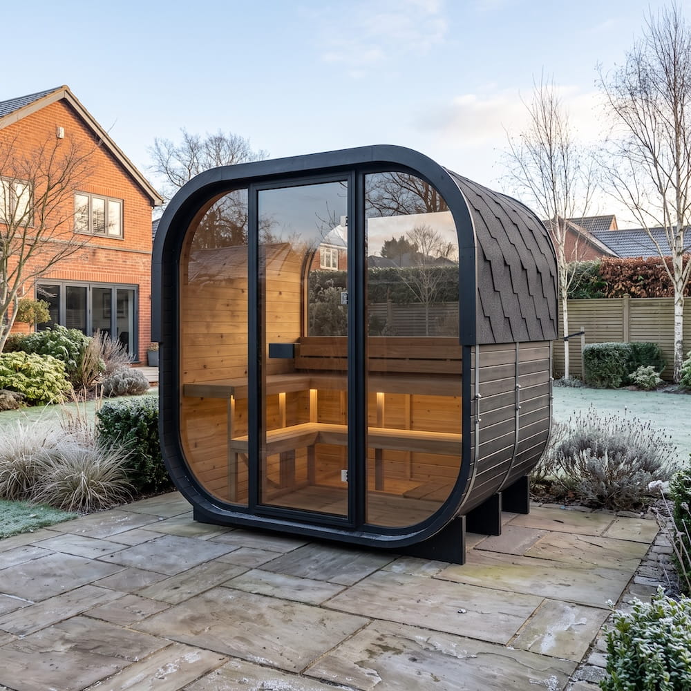 A modern outdoor sauna cabin with a black metal frame, curved glass front, and dark shingled roof, situated on a stone patio in a landscaped garden.