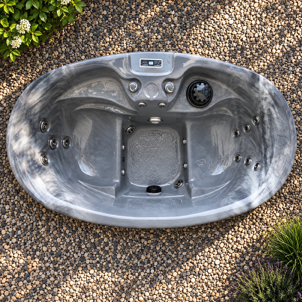 A gray outdoor hot tub with a digital control panel, shown from a top-down view on a bed of small decorative gravel with small plants in the corners.