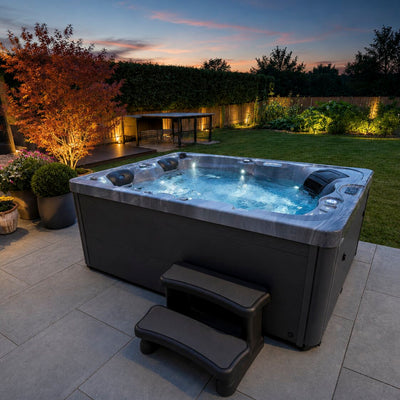An outdoor hot tub with bubbling blue water sits on a paved backyard patio at dusk, with a two-step stair, grassy lawn, trees, and illuminated landscaping in the background.