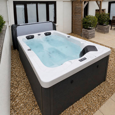 An outdoor rectangular hot tub with a white interior and dark charcoal exterior, filled with bubbling water, set on a gravel patio next to a house.