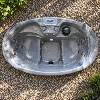 A gray outdoor hot tub with a digital control panel, shown from a top-down view on a bed of small decorative gravel with small plants in the corners.