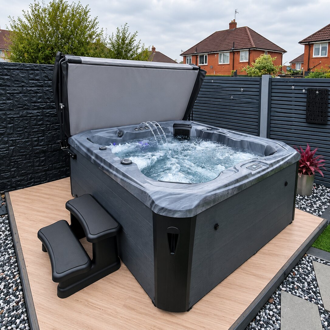 A black hot tub with its lid open, set on a wooden deck in a backyard with a dark gray fence, garden plants, and neighboring houses visible in the background.